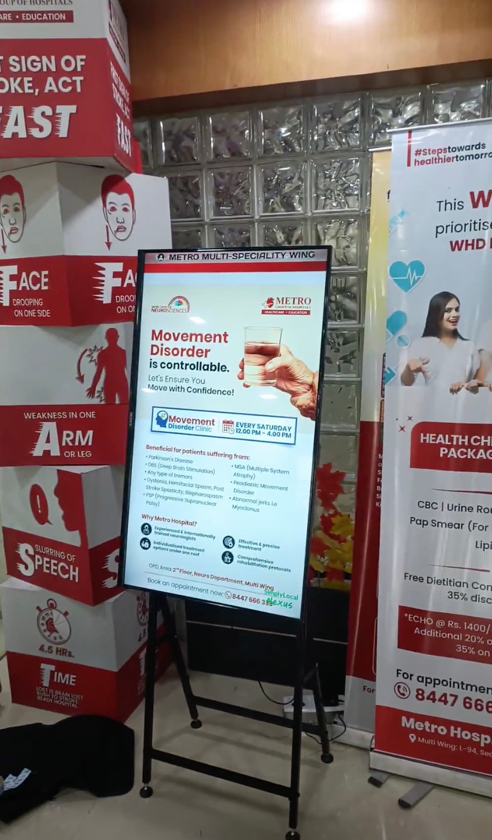 A portrait SLX On Air screen on a stand at Metro Hospital's Multi-Speciality Wing, showing a Movement Disorder Clinic notice